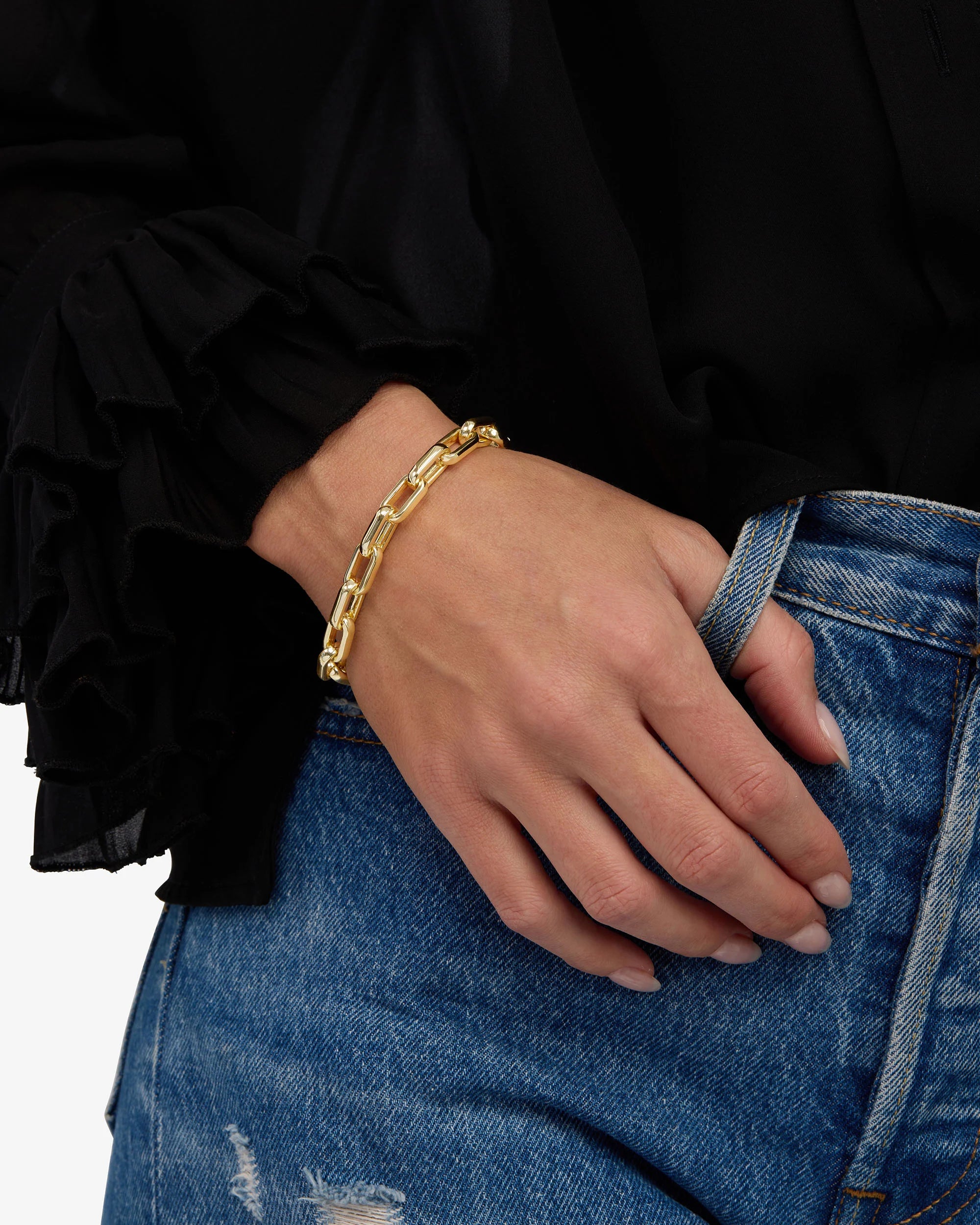A close-up of a person's hand wearing a chunky gold chain link bracelet, styled with a black ruffled blouse and blue distressed denim jeans.