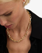 A close-up of a model wearing a chunky gold chain necklace with elongated links, styled with a black collared shirt.