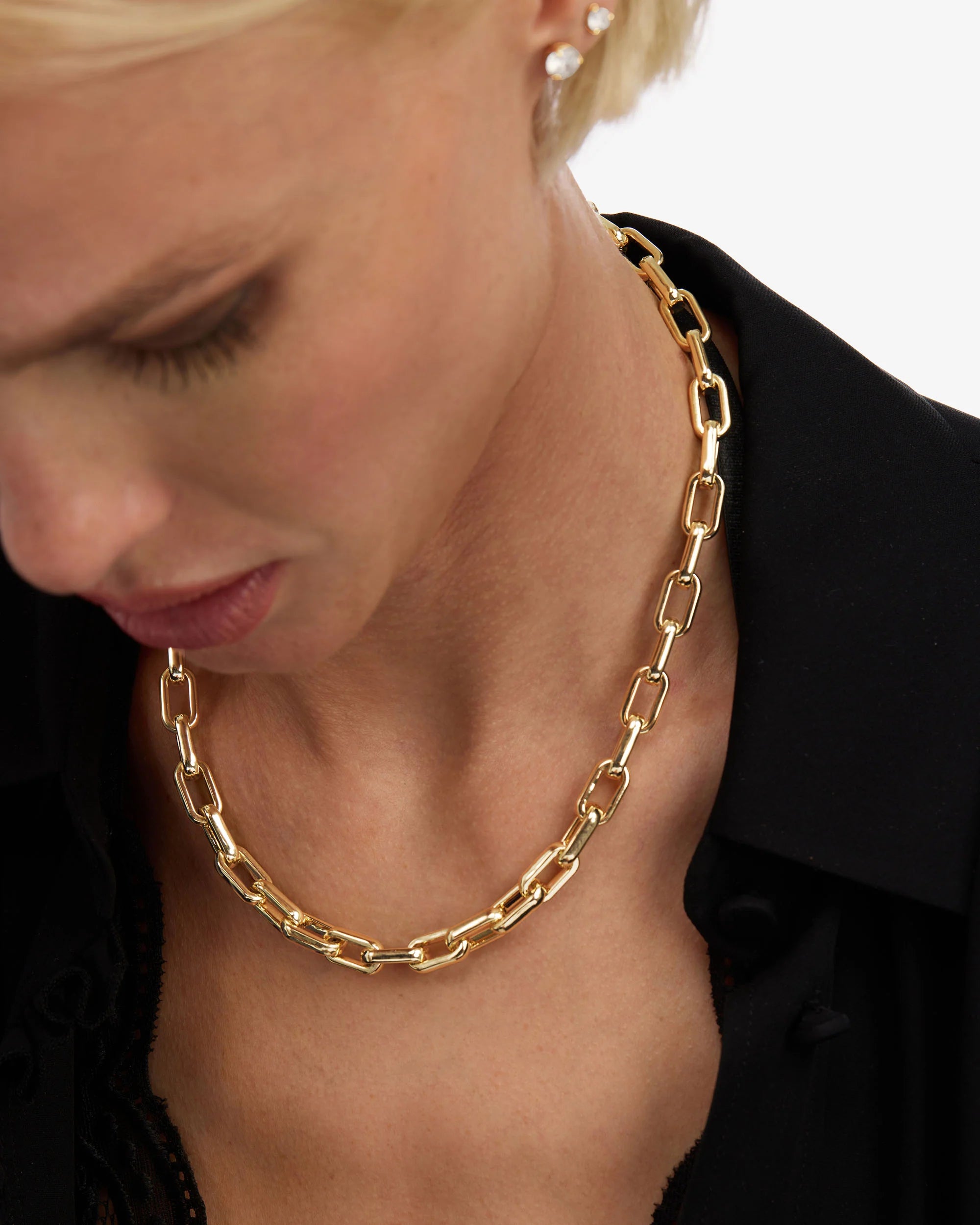 A close-up of a model wearing a chunky gold chain necklace with elongated links, styled with a black collared shirt.