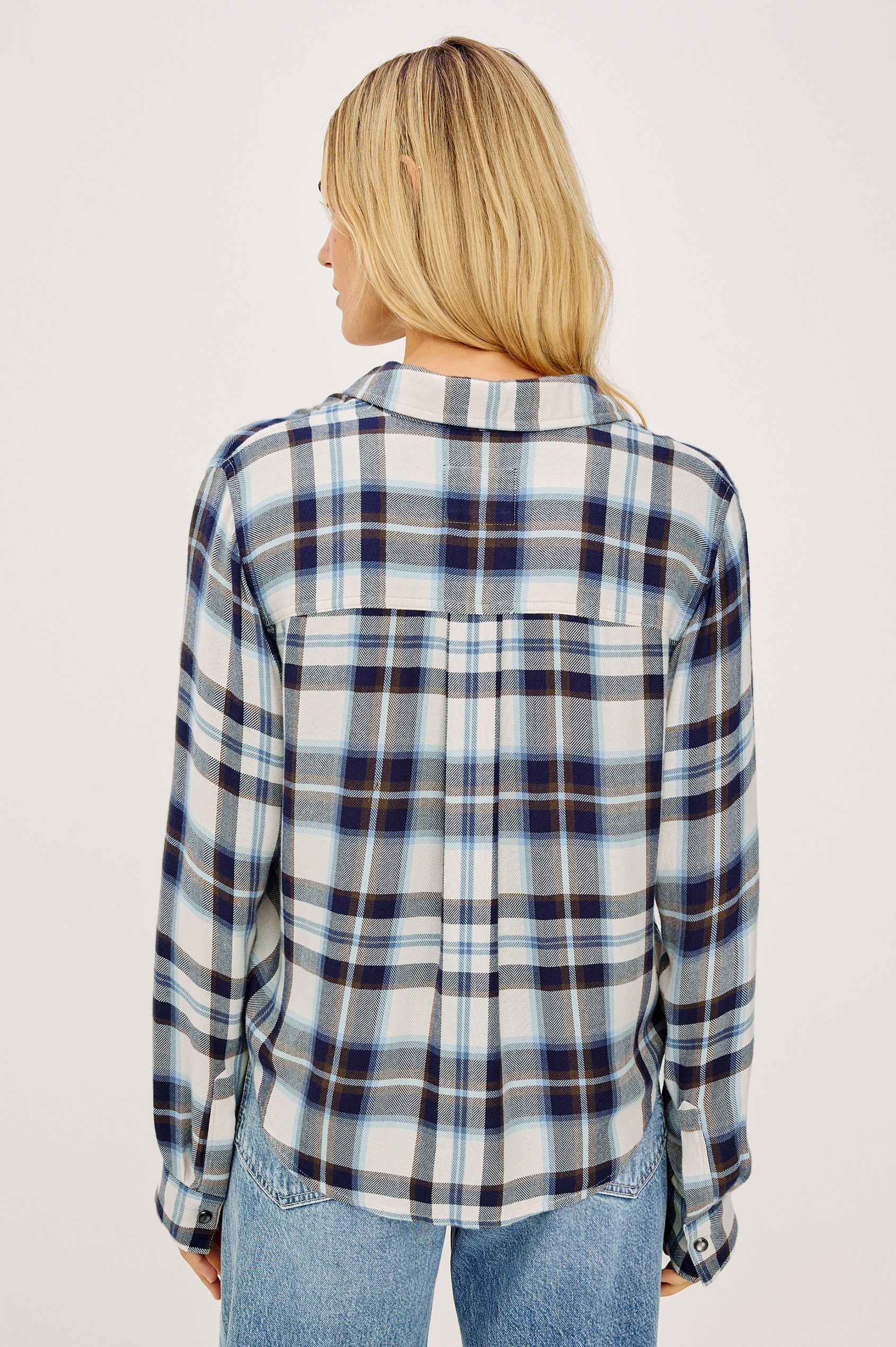 A back view of a woman wearing a long-sleeve button-up plaid shirt in blue, white, and navy, paired with light-wash blue jeans, photographed against a light gray studio background.
