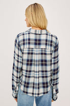A back view of a woman wearing a long-sleeve button-up plaid shirt in blue, white, and navy, paired with light-wash blue jeans, photographed against a light gray studio background.