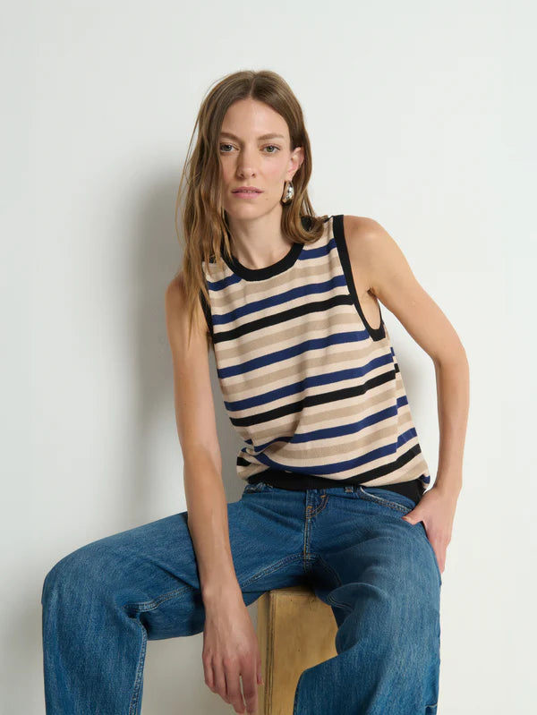 Woman wearing a striped tank top and jeans sitting on a wooden stool against a plain background