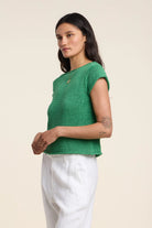 Woman wearing a green knitted top and white pants against a beige background