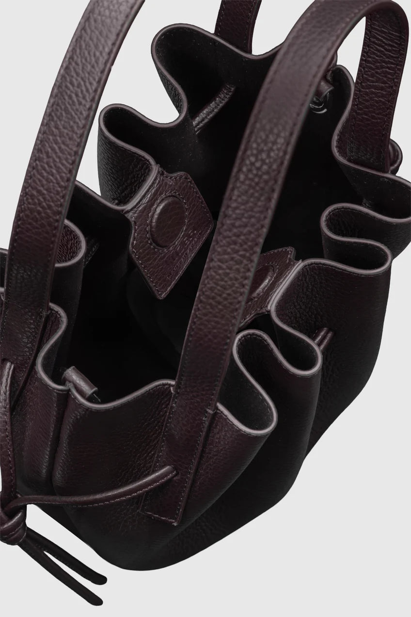 A close-up of a dark brown pebbled leather bucket bag with ruffled trim and drawstring ties, shown against a light gray background.
