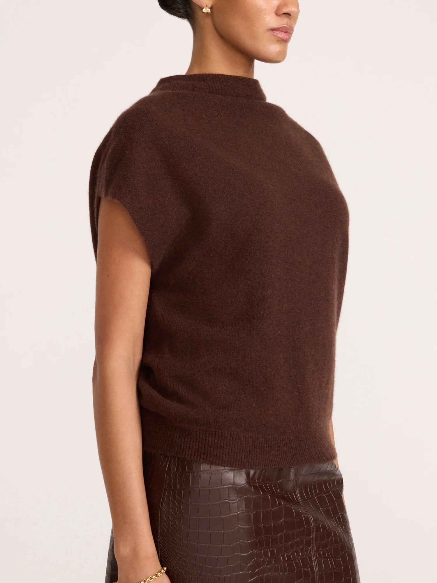 A side-profile view of a model wearing a dark brown sleeveless mock-neck knit sweater and a dark brown croc-embossed skirt, against a plain off-white background.