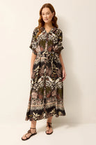 A woman modeling a short-sleeve midi dress with a botanical and tribal pattern in olive green, black, and beige, paired with black strappy sandals against a plain off-white background.