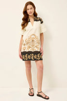 Woman wearing a white dress with black and gold embroidery on a plain background