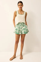Woman wearing a white sleeveless top and green floral shorts on a plain background