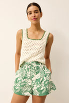 Woman wearing a white top with green trim and green floral shorts on a plain background
