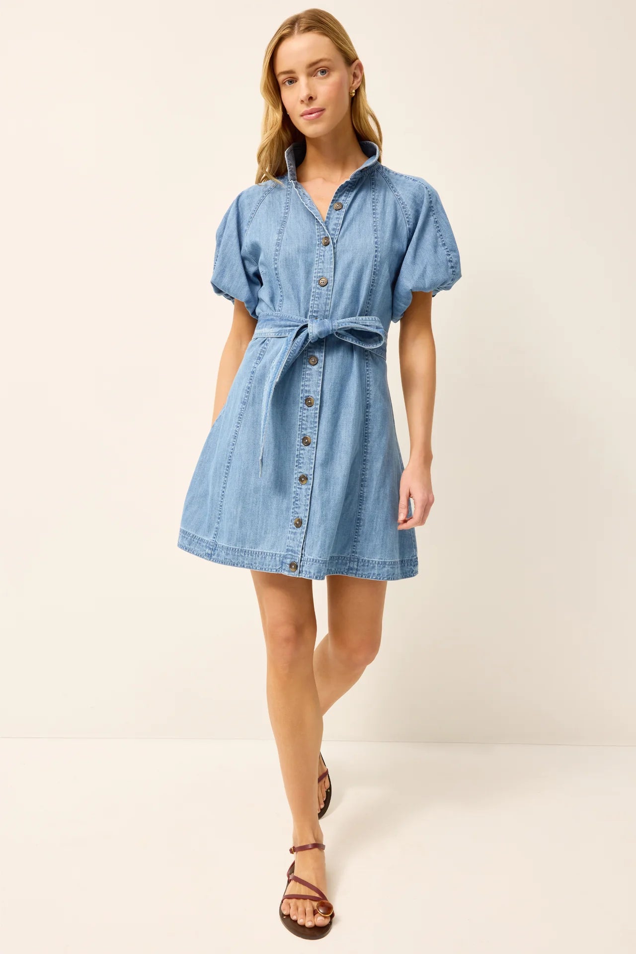 A woman modeling a light blue denim shirt dress with short puff sleeves, a button-front closure, and a self-tie belt at the waist, paired with brown strappy sandals against a plain cream studio background.