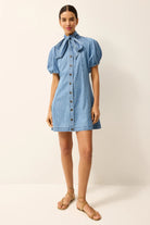 A model wearing a light blue denim mini dress with puff sleeves, a tie-neck bow, and a button-down front, paired with beige strappy sandals against a plain cream studio background.