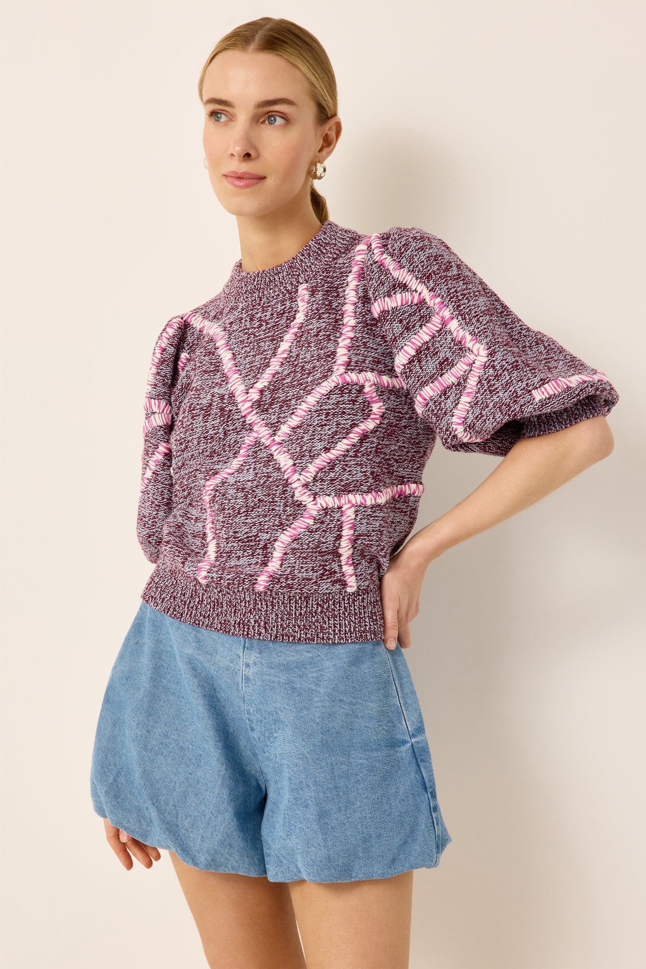 A woman modeling a short-sleeve knit sweater with a marled purple and pink pattern, paired with light blue denim shorts, against a plain beige background.