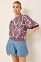 A woman modeling a short-sleeve knit sweater with a marled purple and pink pattern, paired with light blue denim shorts, against a plain beige background.
