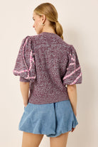 Woman wearing a maroon and pink top with puff sleeves and a blue skirt on a beige background