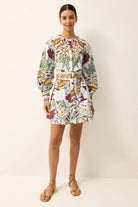 A woman modeling a long-sleeve mini dress with a white base and a colorful botanical print in green, purple, yellow, and orange, featuring a matching fabric belt. She is wearing tan strappy sandals against a plain beige studio background.
