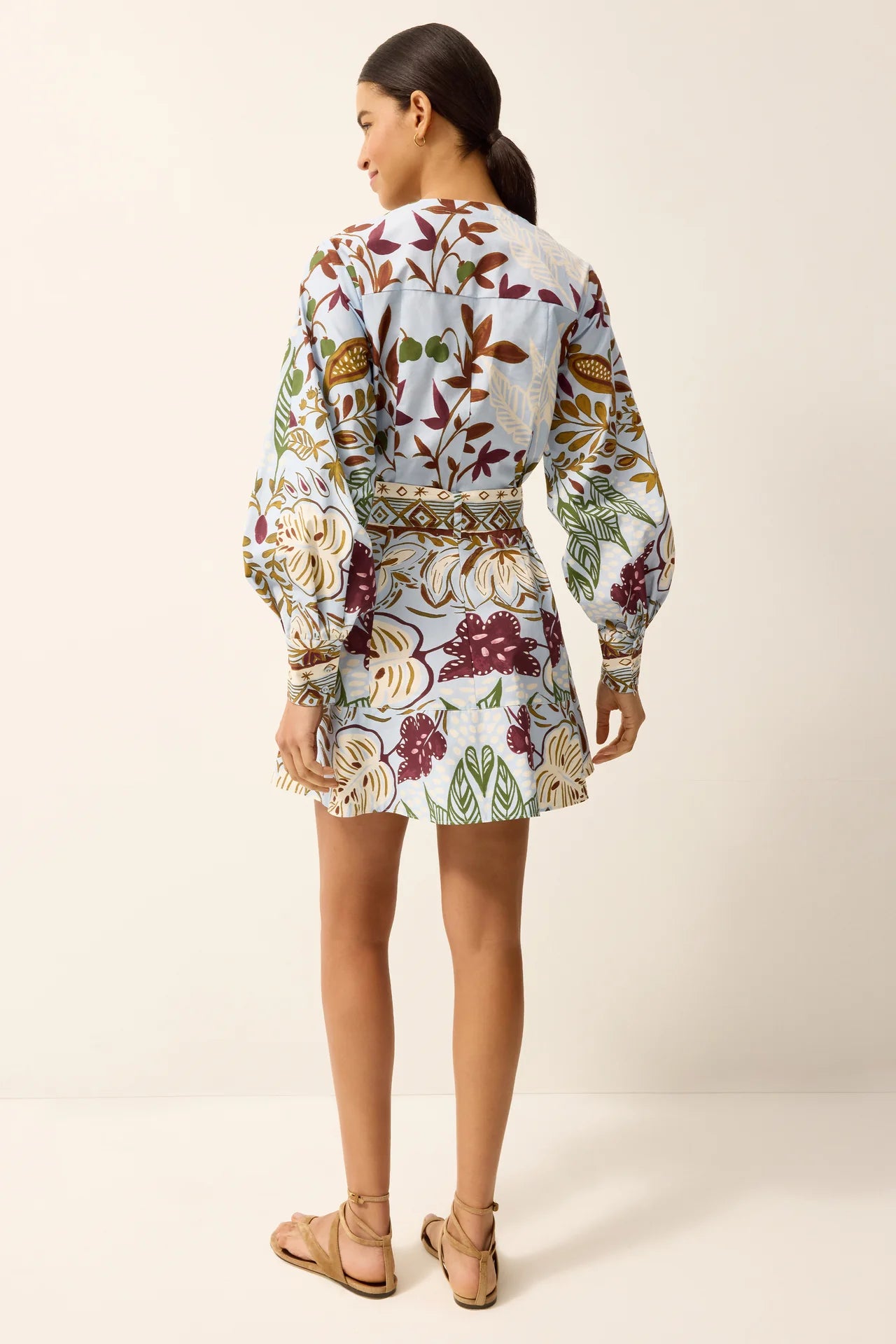 A woman shown from the back wearing a long-sleeve mini dress with a colorful floral and botanical print on a light background, paired with tan flat sandals, against a plain beige studio backdrop.