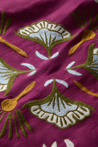 A close-up of a magenta fabric with embroidered botanical motifs in olive green, cream, and mustard yellow.