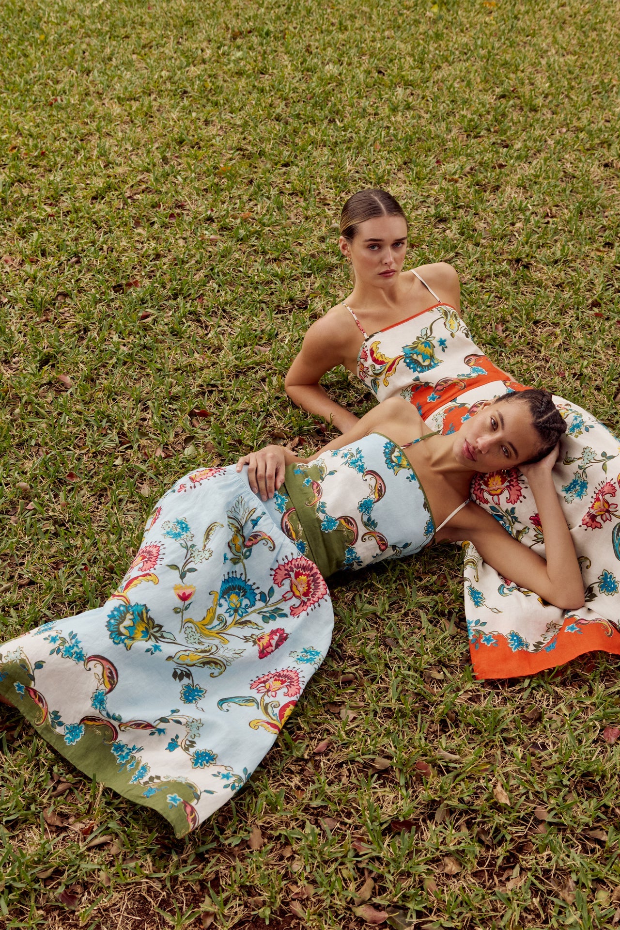 Two women lying on grass wearing colorful floral dresses.