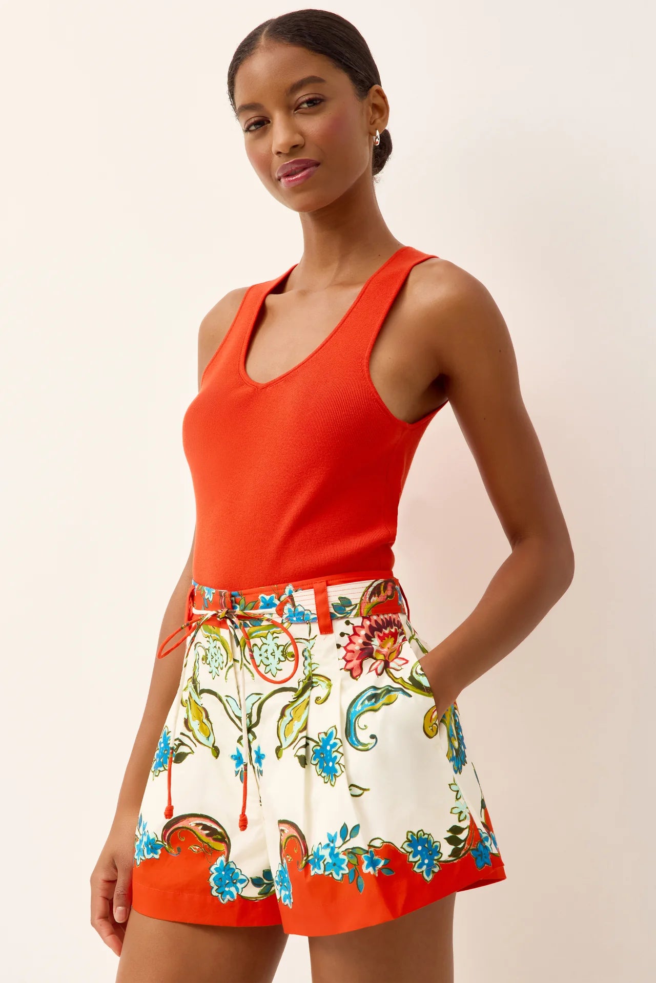 Woman wearing a bright orange sleeveless top and colorful floral skirt on a plain background