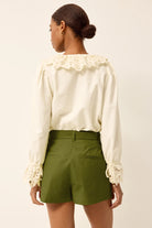 Woman wearing a white blouse with lace details and green shorts on a beige background
