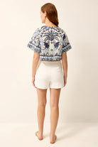Woman wearing a patterned top and white shorts on a plain background
