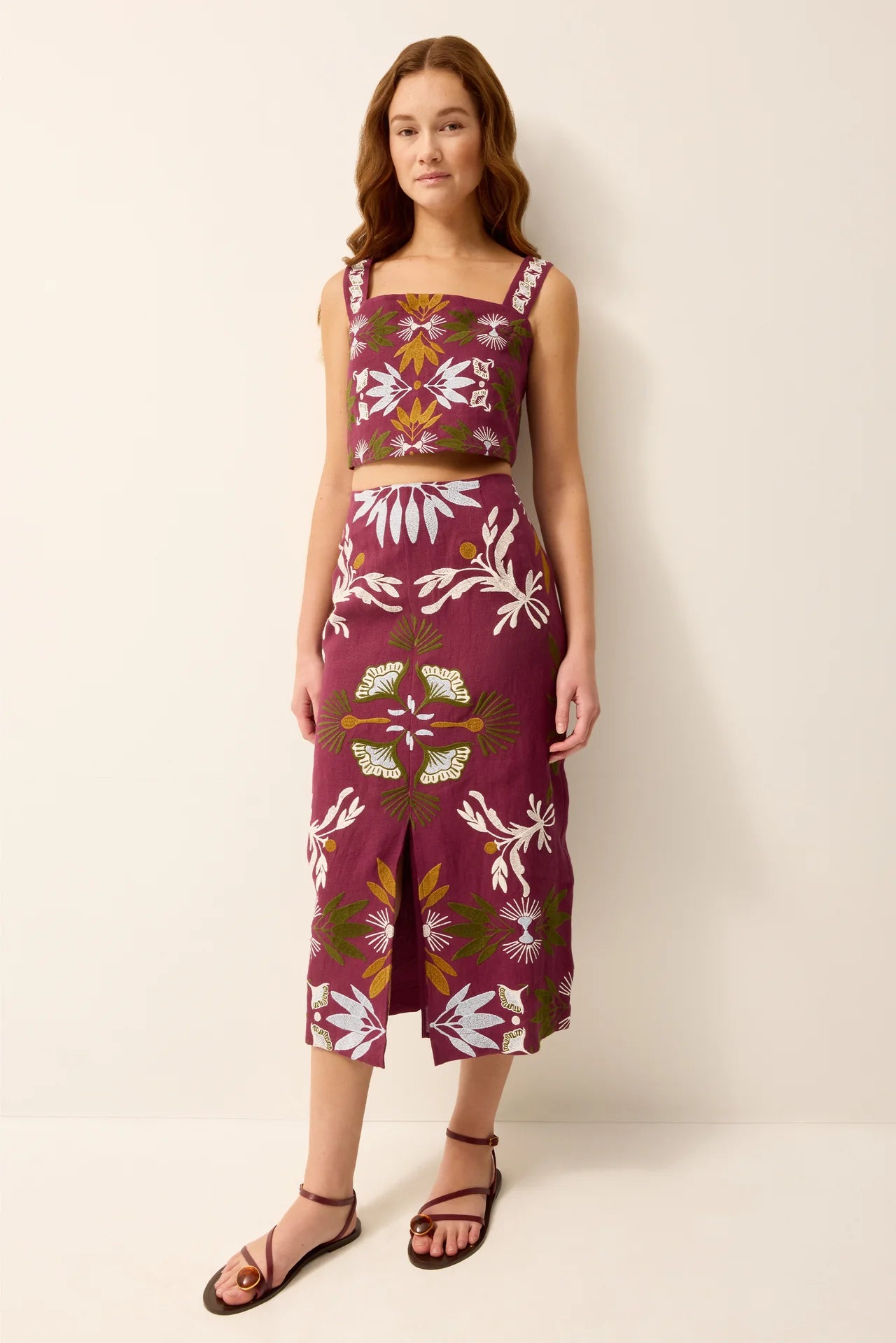 A woman modeling a matching two-piece set consisting of a sleeveless crop top and a midi skirt, both in a deep burgundy color with a white, green, and gold botanical pattern, set against a plain beige studio background.