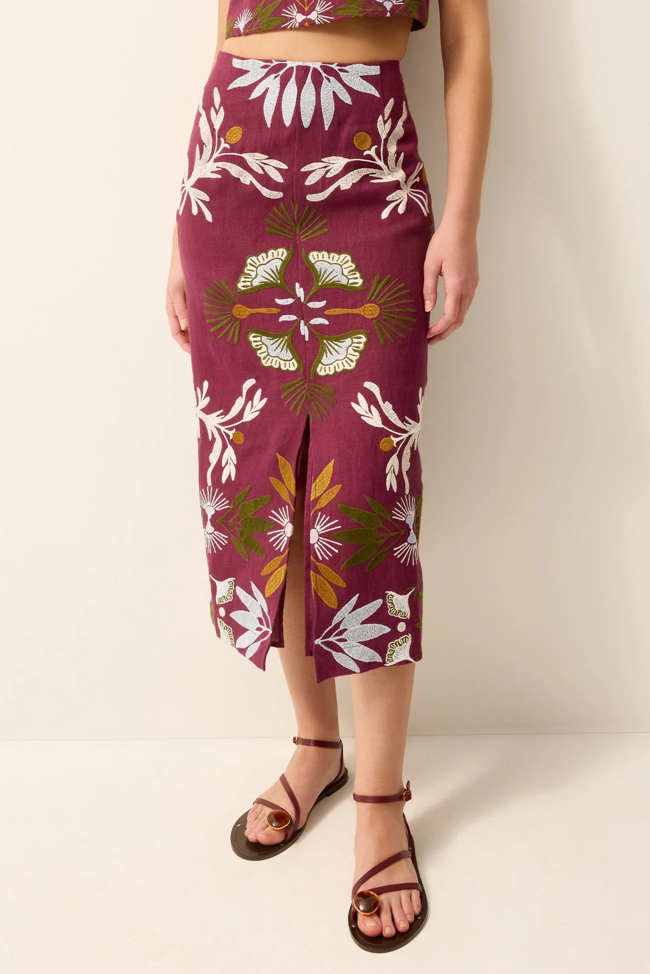 A model wearing a high-waisted, knee-length skirt with a floral print in burgundy, white, and olive green, paired with brown strappy sandals against a plain beige studio background.