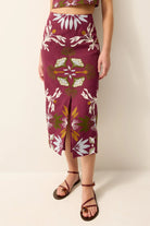 A model wearing a high-waisted, knee-length skirt with a floral print in burgundy, white, and olive green, paired with brown strappy sandals against a plain beige studio background.
