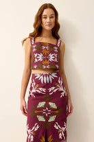 A woman modeling a matching two-piece set featuring a sleeveless crop top and a high-waisted skirt, both in a deep burgundy color with a white, olive green, and mustard yellow botanical pattern, set against a plain off-white background.