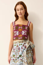 A woman modeling a sleeveless top and matching patterned skirt set against a plain beige studio background. The top has a deep magenta-burgundy base with white, olive green, and mustard yellow floral and geometric motifs.