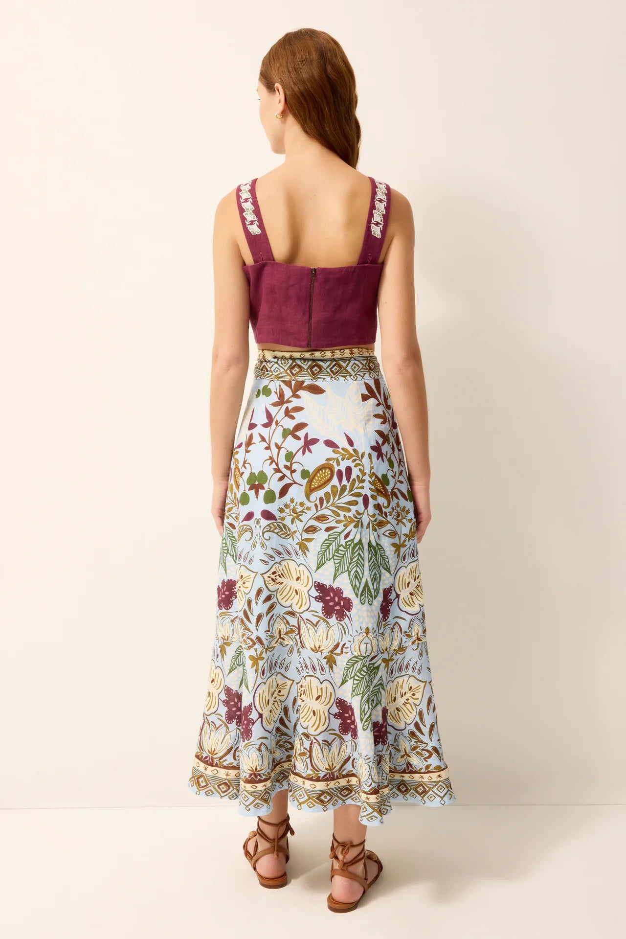 A woman shown from the back wearing a sleeveless maxi dress with a deep burgundy scoop neck and a long white skirt featuring a colorful floral and botanical print, paired with brown strappy sandals against a plain beige studio background.