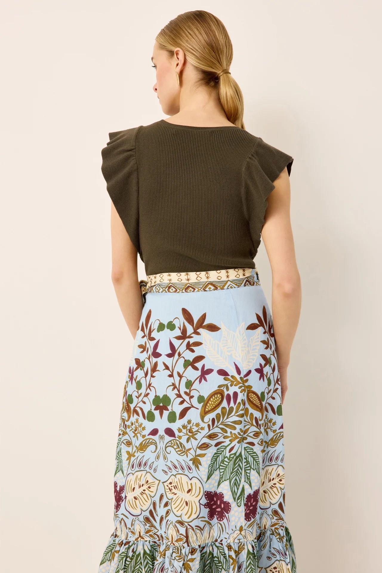 A woman shown from the back wearing a dark olive green ruffled short-sleeve top and a light blue floral print skirt with a patterned belt, against a plain beige background.