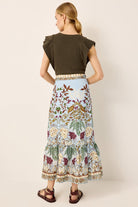 A woman shown from the back wearing a dark olive green short-sleeve top and a long light blue tiered skirt with a detailed floral and bird print, paired with brown sandals, against a solid beige background.