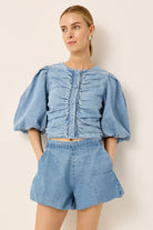 A woman modeling a light blue denim two-piece set consisting of a cropped top with dramatic puff sleeves and front pleating, paired with matching high-waisted shorts, against a plain beige background.