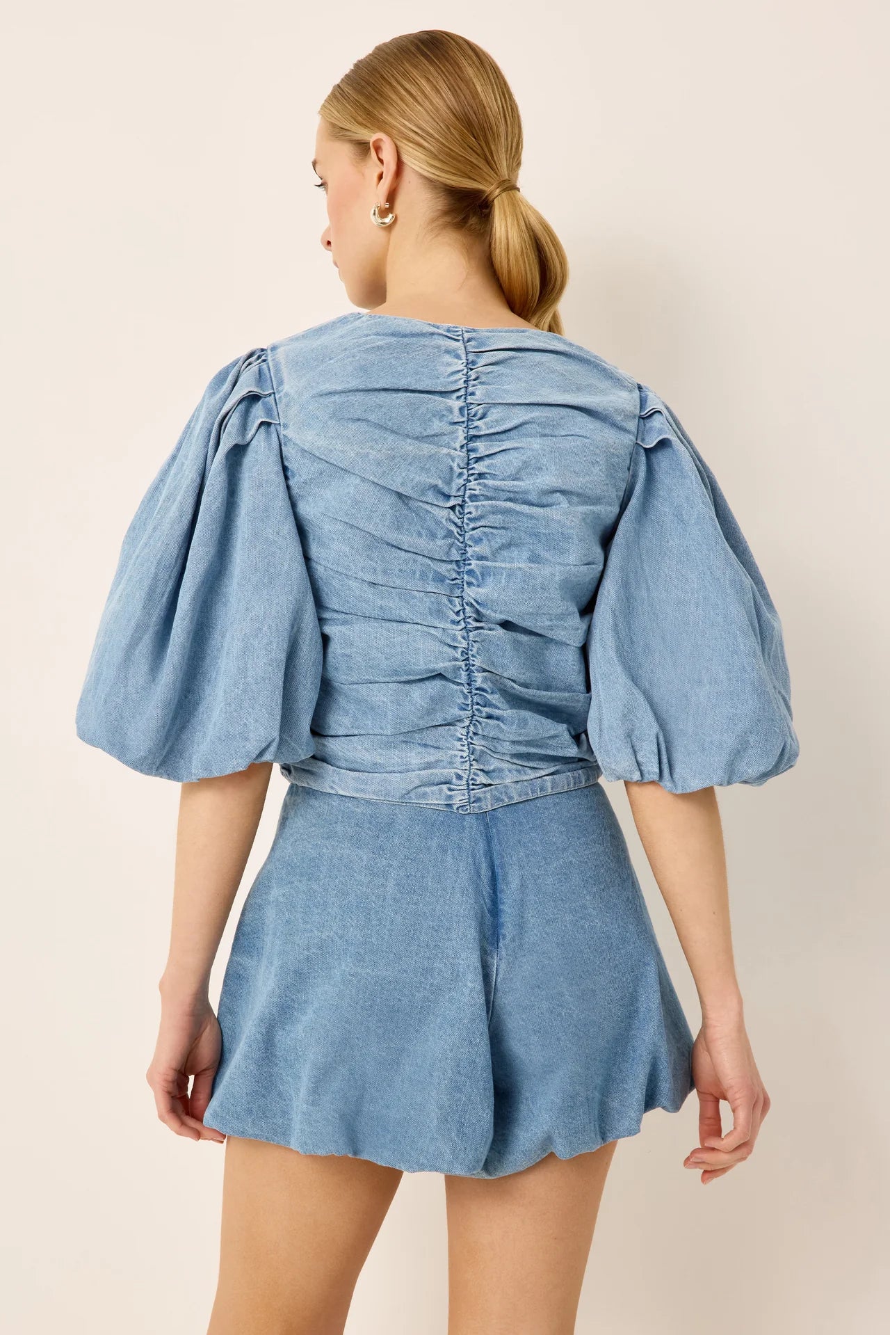A woman shown from the back wearing a light blue denim romper with puffed sleeves and a ruched back, against a plain beige studio background.