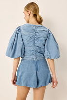 A woman shown from the back wearing a light blue denim romper with puffed sleeves and a ruched back, against a plain beige studio background.