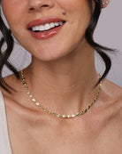 A close-up of a woman wearing a delicate gold chain necklace with small, flat, oval-cut gold stones set along the chain, paired with a white top against a neutral light gray background.