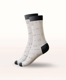 A pair of crew socks with a white base, black cuffs, heels, and toes, and a minimalist black line-art face pattern, shown floating against a light beige gradient background.