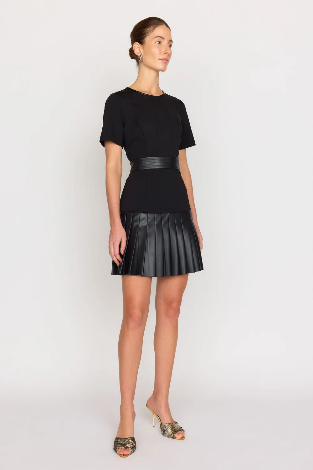 A woman modeling a black short-sleeve mini dress with a wide waistband and a pleated skirt, paired with patterned high-heeled sandals, against a plain light gray studio background.