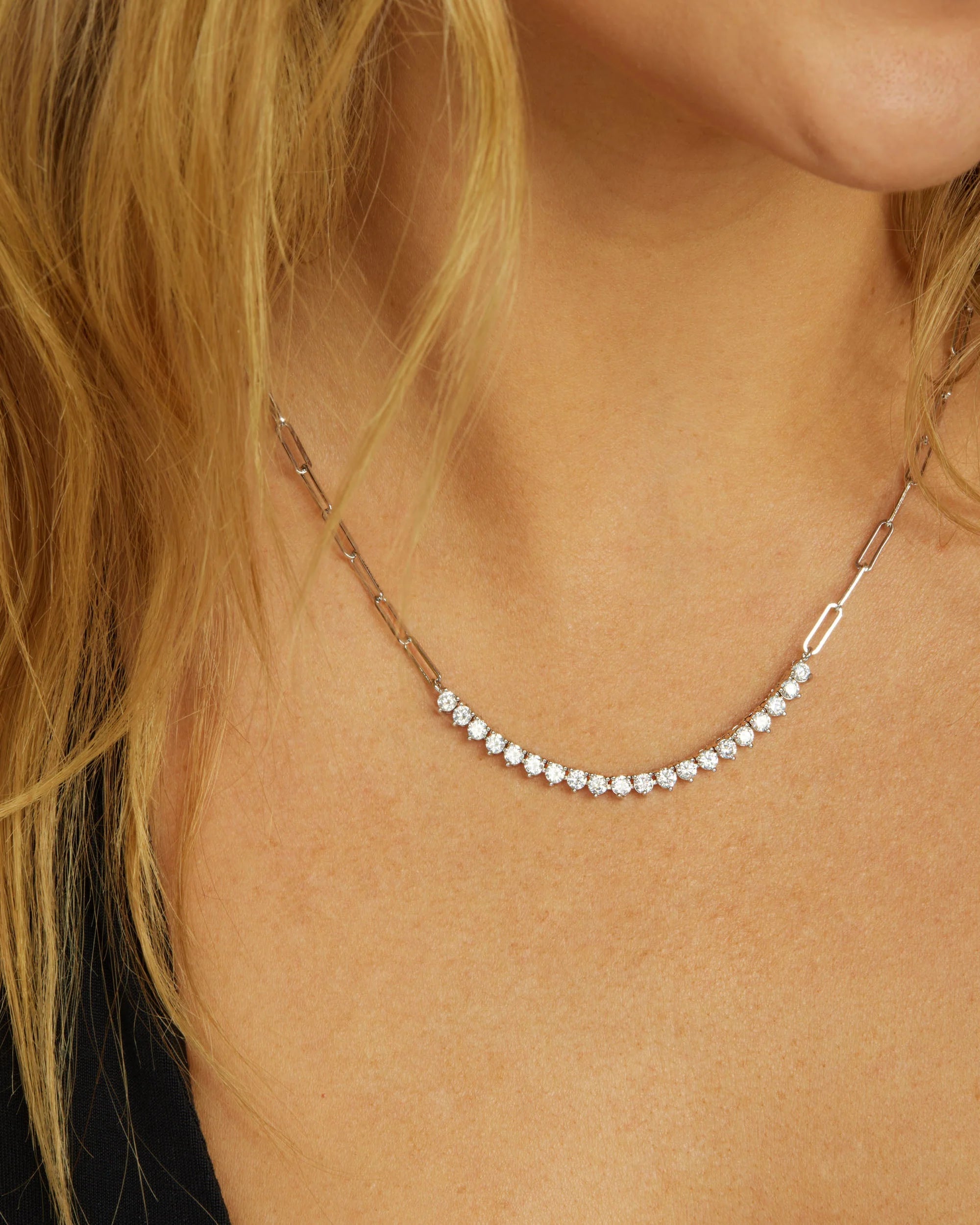 A close-up of a woman's neck and collarbone wearing a delicate silver tennis necklace with a paperclip clasp.
