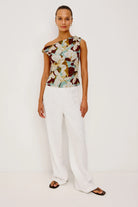Woman wearing a floral one-shoulder top and white pants on a plain background