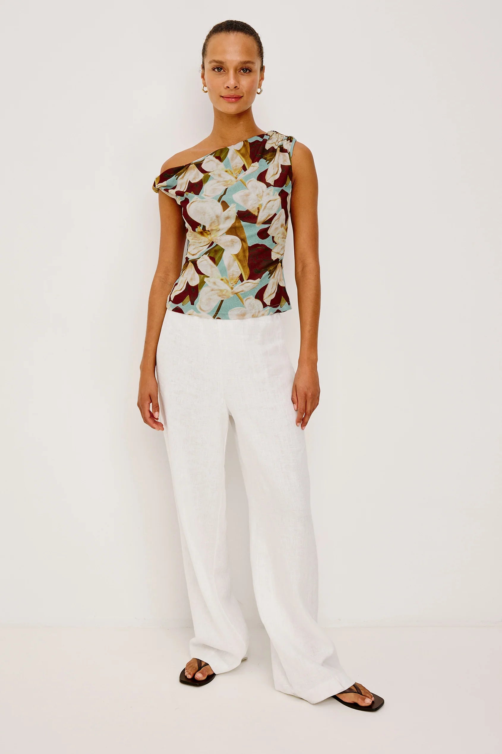 Woman wearing a floral one-shoulder top and white pants on a plain background