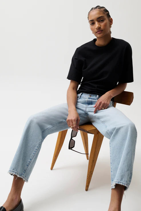 Person wearing a black t-shirt and light blue jeans sitting on a wooden chair against a white background