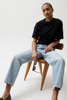 Person wearing a black t-shirt and light blue jeans sitting on a wooden chair against a white background