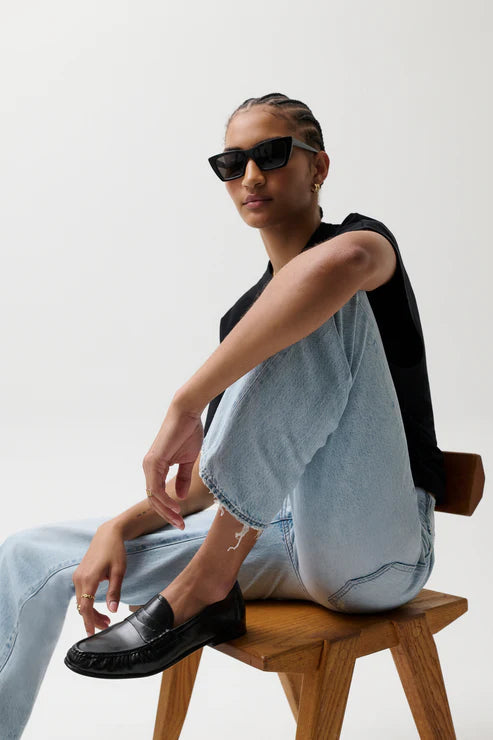 Person wearing sunglasses and light blue jeans sitting on a wooden stool against a white background