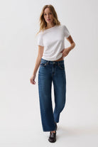 A woman modeling a pair of wide-leg blue jeans with a white t-shirt and dark brown leather loafers, standing against a plain white studio background.