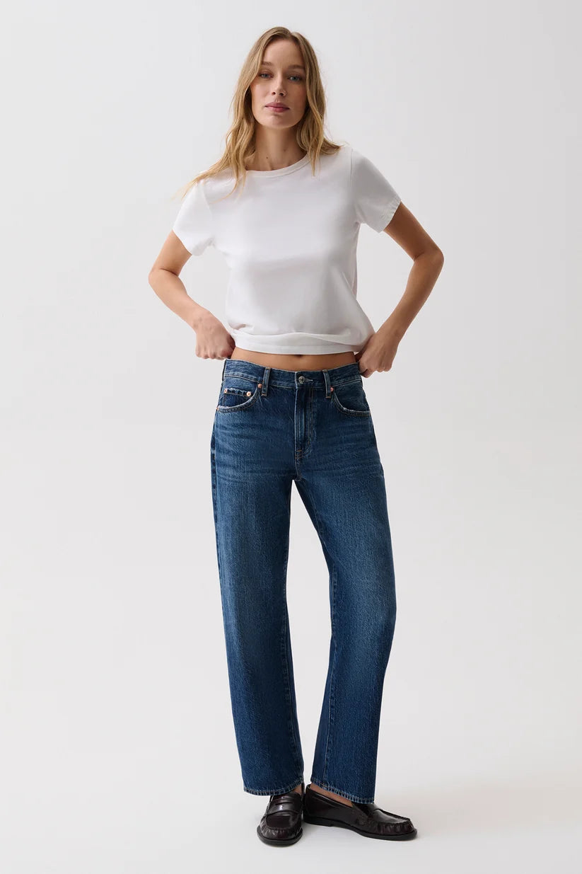 A woman modeling a pair of dark blue straight-leg jeans with a white short-sleeve crop top and black loafers, standing against a light gray studio background.