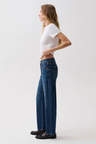 A woman in a white t-shirt and blue wide-leg jeans stands in profile against a plain light gray studio background.