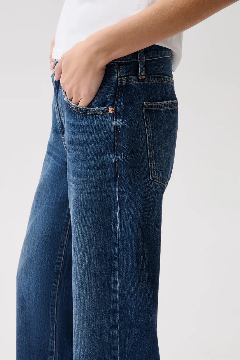 A close-up of a person wearing dark blue wide-leg denim jeans with a white top, showing the side pocket and front seam details against a light gray background.