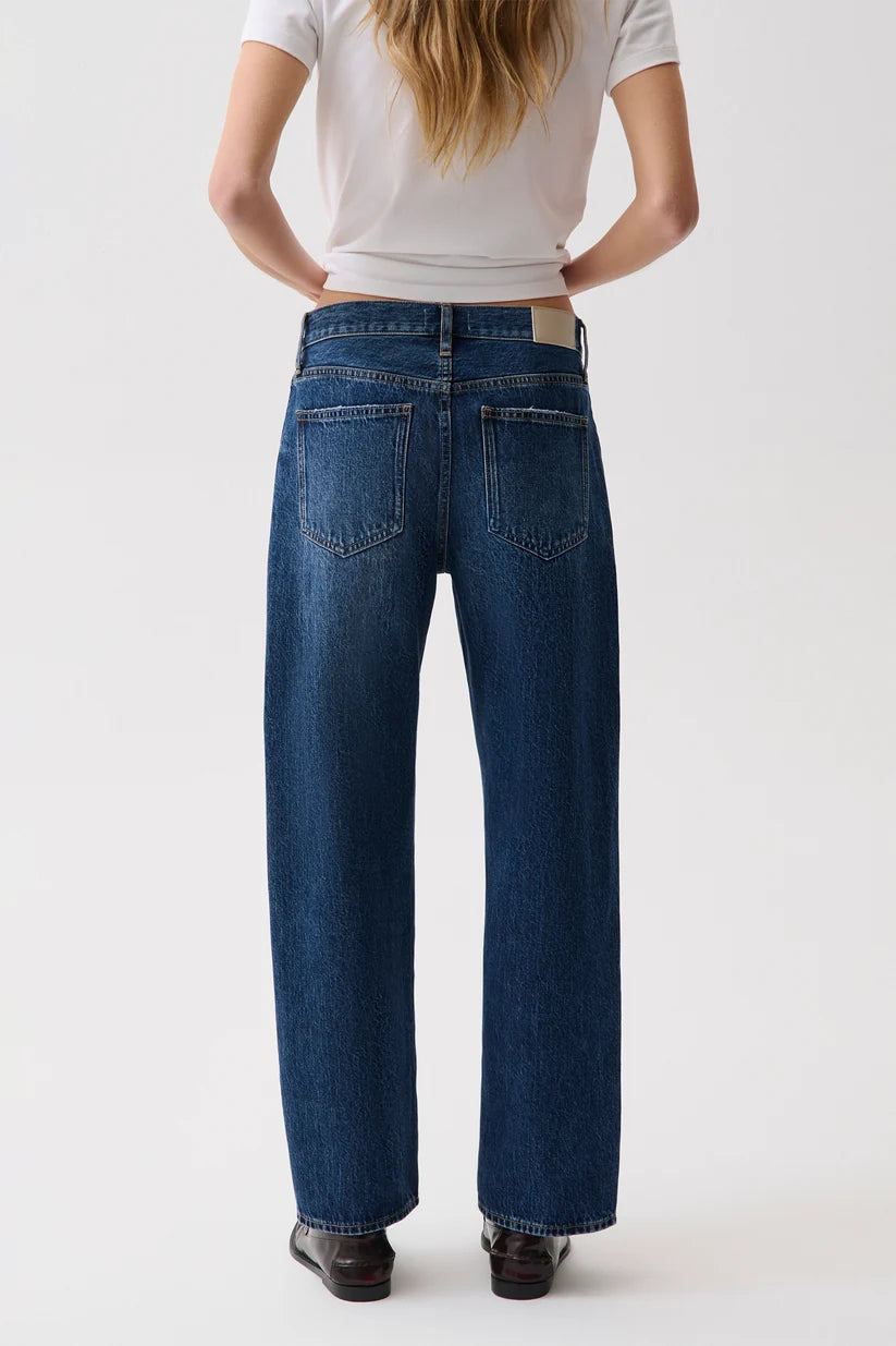 A model viewed from the back wearing a pair of dark blue wide-leg jeans, a white t-shirt, and dark brown shoes, set against a plain white studio background.